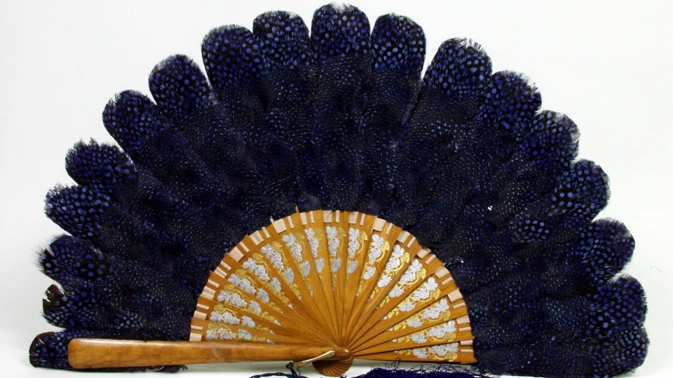 Vějíř skládací s peřím perličky africké. Dřevěná, lakovaná žebra (17 + 2) jsou zdobeny rytým, zlaceným a stříbřeným stylizovaným rostlinným ornamentem