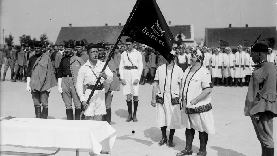 Fotografie zachycující rozvinutí sokolského praporu a sokolský průvod. Na vlajkách a dámském oblečení je dobře patrná svérázová výšivka. Balley, 16. 6. 1929